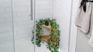 A shower with white tiled walls, a glass door, a towel rack with a towel, and a small shelf with greenery.