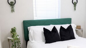 A neatly made bed with white bedding and black pillows, a green upholstered headboard, and a window with blinds.
