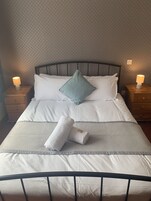 Una cama bien tendida con sábanas blancas y una única almohada color aguamarina, flanqueada por dos lámparas de noche sobre mesitas de madera.