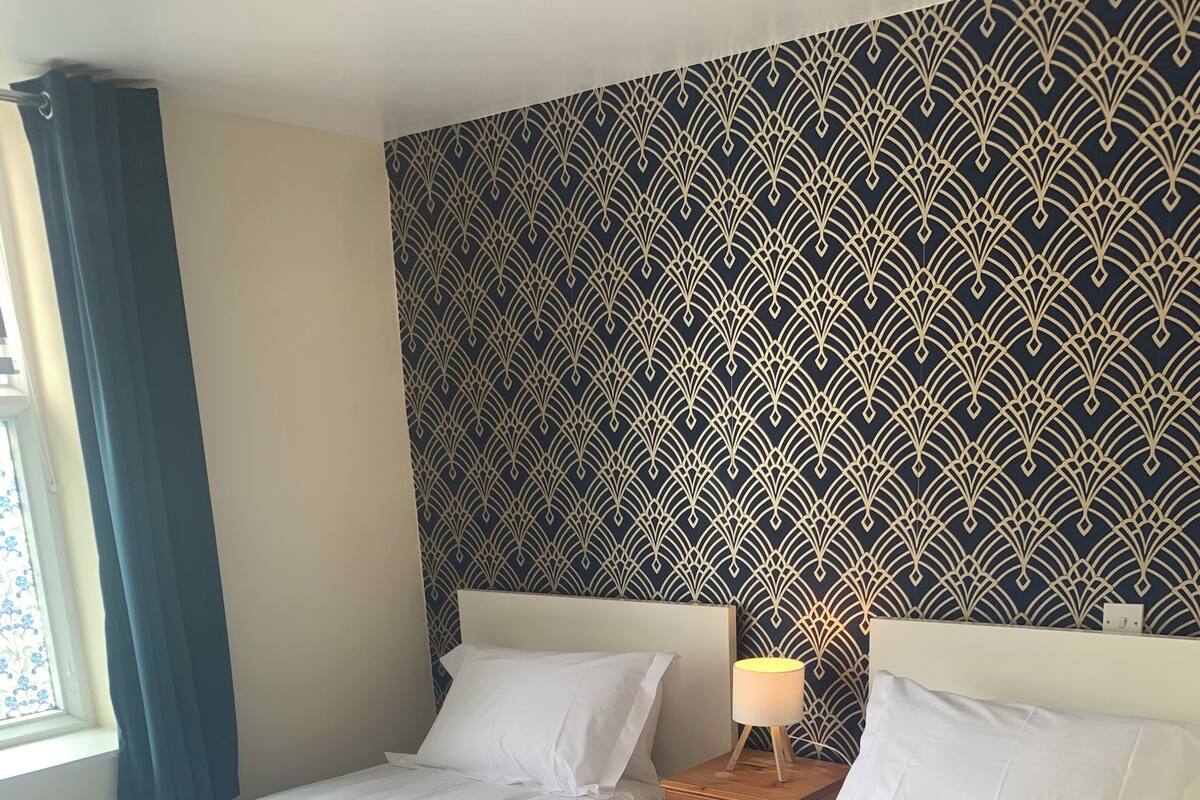 A hotel room with two beds, a patterned wallpaper, a window with curtains, and a bedside lamp.