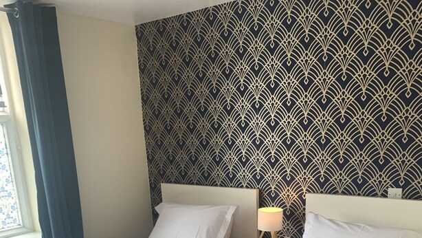 Habitación de hotel con dos camas, un papel tapiz estampado, una mesita de noche con lámpara y una ventana con cortinas.