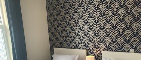 Une chambre d’hôtel avec deux lits, un papier peint à motifs, une table de chevet avec une lampe et une fenêtre avec des rideaux.