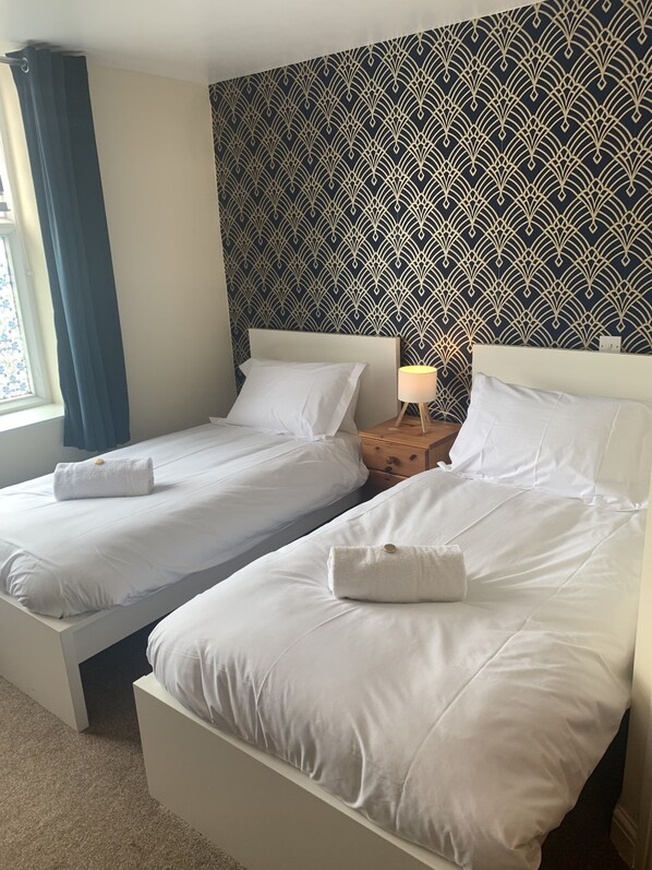 A hotel room with two beds, a patterned wallpaper, a bedside table with a lamp, and a window with curtains.