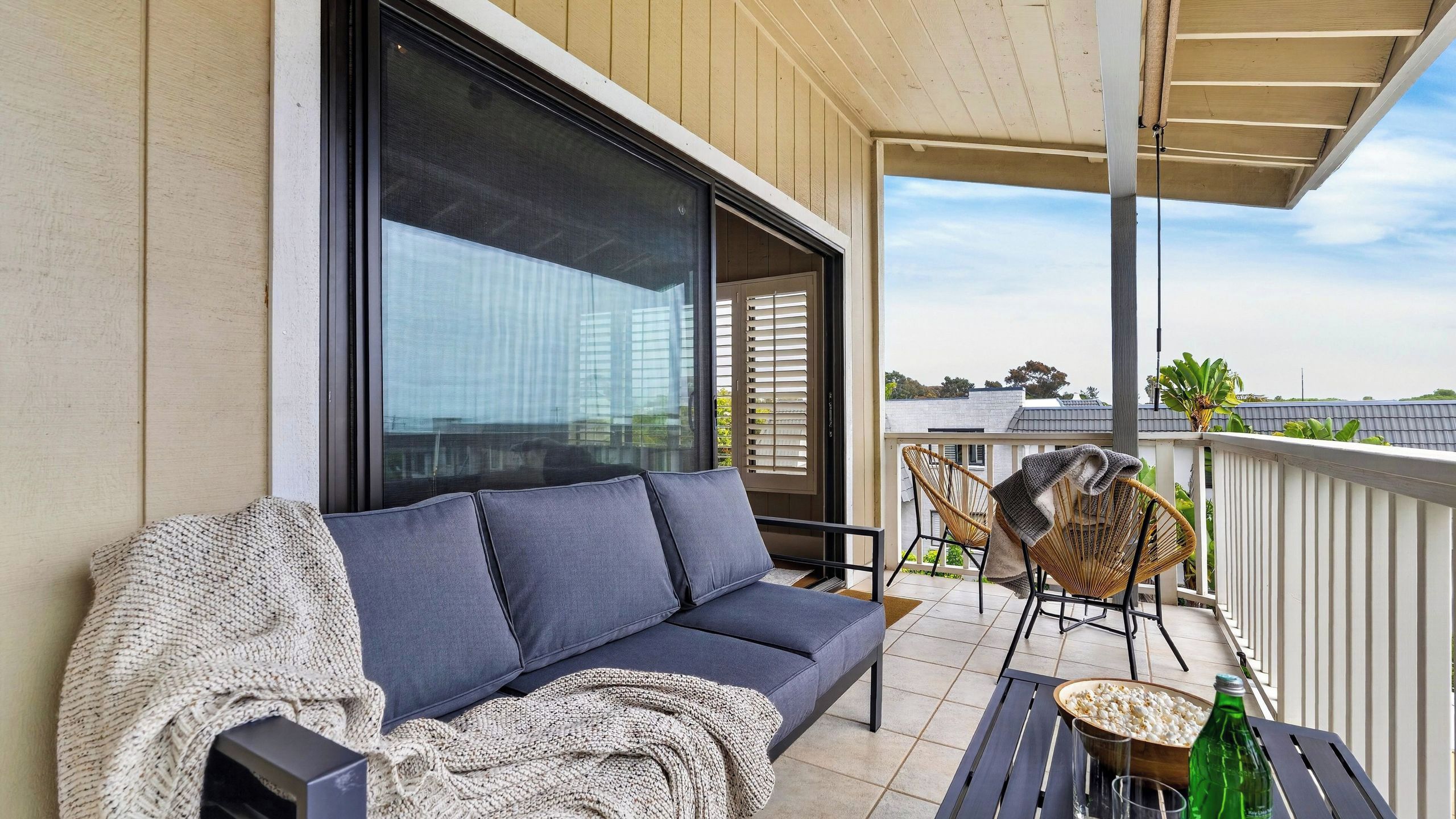 A balcony with a sofa, two wicker chairs, a table with popcorn and a bottle, and a view of buildings and trees.