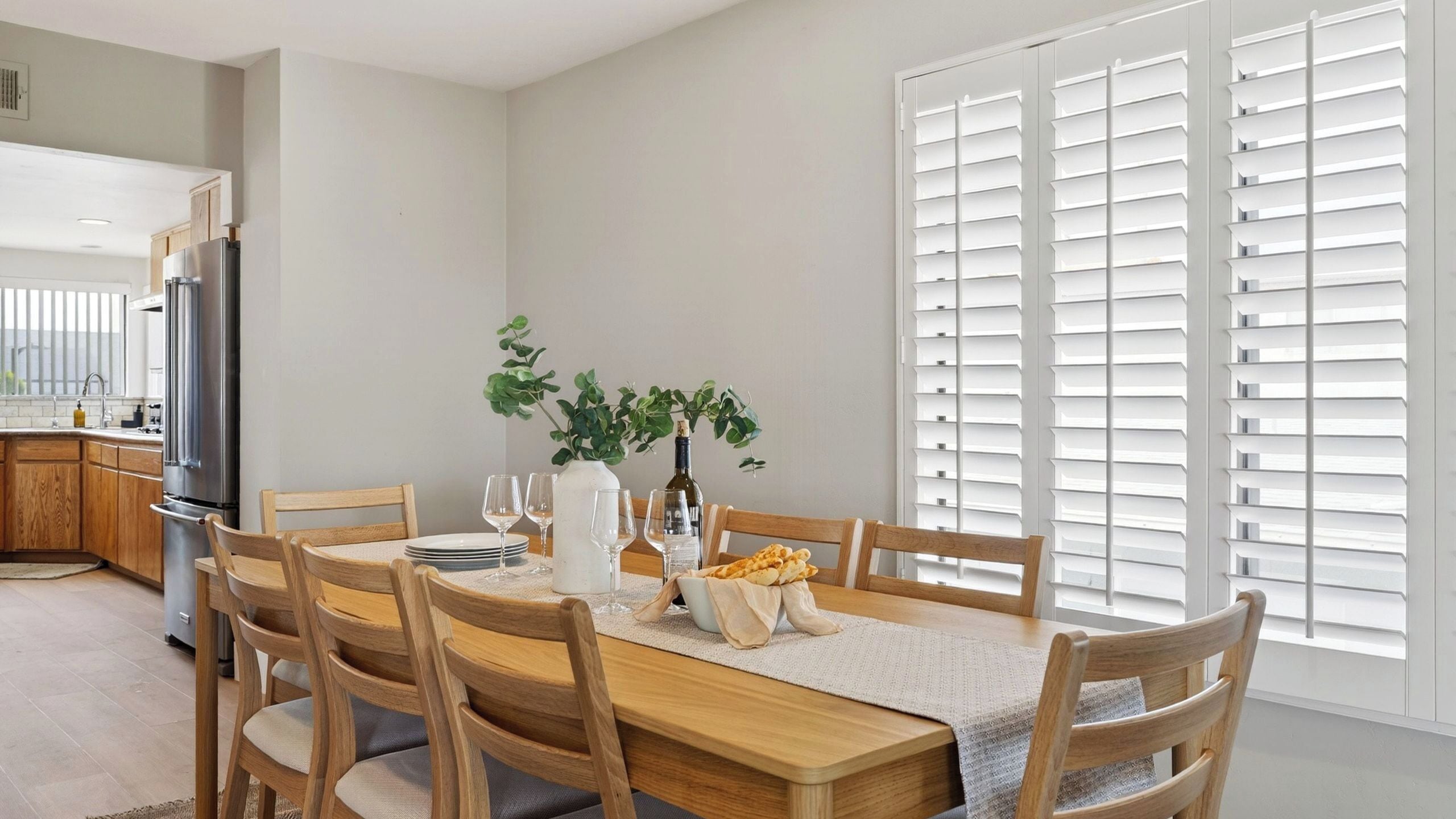 A dining area with a wooden table, chairs, a vase with plants, wine glasses, and a bottle.