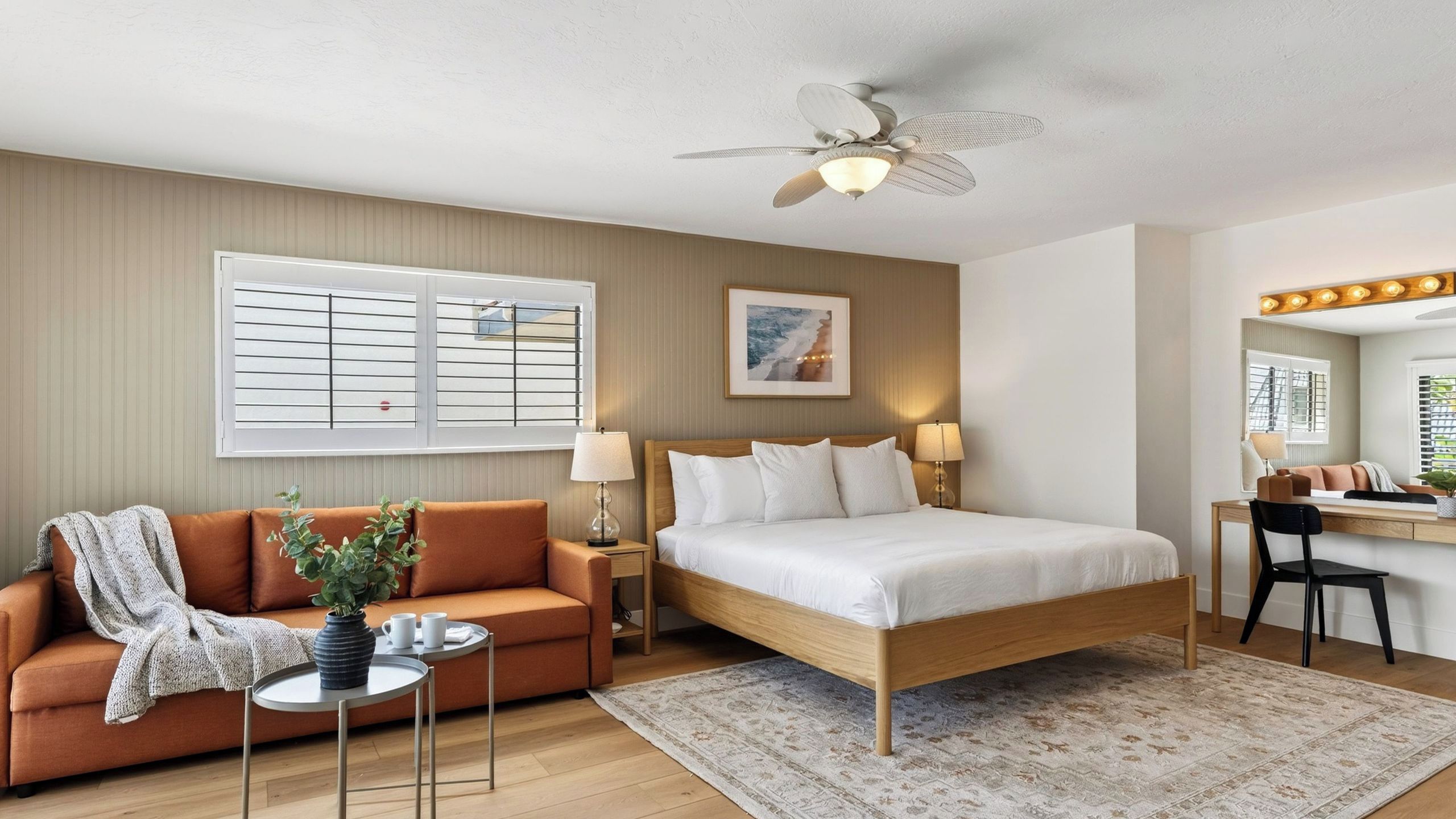A hotel room with a bed, a sofa, a desk, a chair, a ceiling fan, a window with blinds, and a framed picture on the wall.