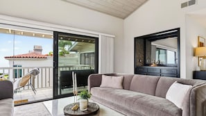 A modern living room with a grey sofa, a round coffee table, a glass door leading to a balcony, and a large mirror on the wall.