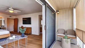 A bedroom with a bed, a small table with a vase, a ceiling fan, and a sliding glass door leading to a balcony with wicker furniture.