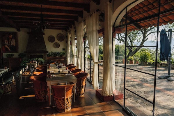 A restaurant with a long table, chairs, and a view through large windows.