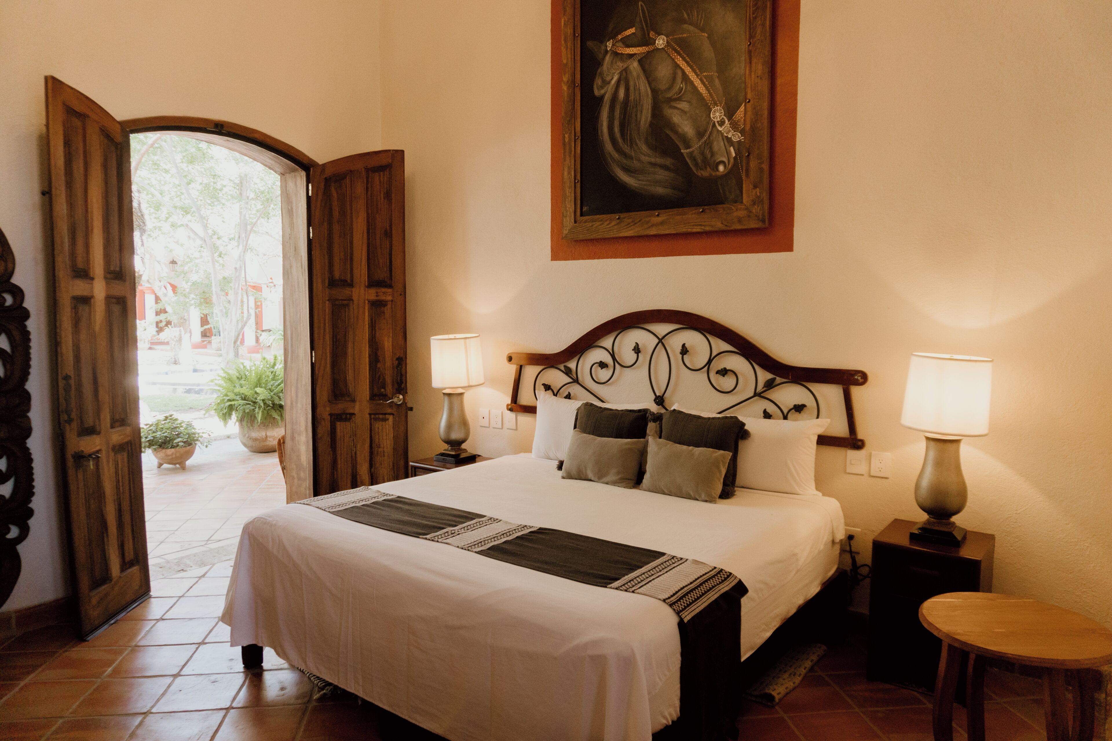 A hotel room with a bed, two bedside lamps, a painting on the wall, and an open door leading to a courtyard.
