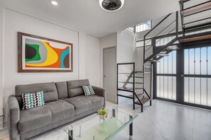 A modern living room with a grey sofa, a glass coffee table, a colorful abstract painting, a staircase, and large windows. - Loft Close to the Ocean With Rooftop Infinity Pool, Balcony & gym - Dog-friendly (Cabo San Lucas)