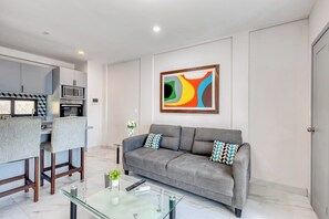 A modern living room with a grey sofa, a glass coffee table, and a kitchen area in the background. - Loft Close to the Ocean With Rooftop Infinity Pool, Balcony & gym - Dog-friendly (Cabo San Lucas)