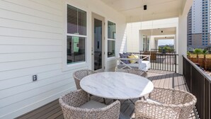 A covered outdoor area with a round table and wicker chairs, a balcony with a view of a building, and a door leading to an interior space. - Steps to Beach! Heated Pool & Modern Luxury (gulf shores)