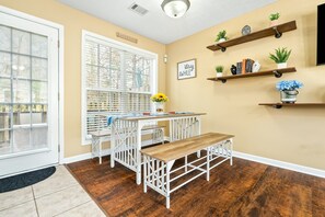 Un coin repas avec une table et un banc blancs, une fenêtre avec des stores et des étagères murales ornées de plantes et de livres.