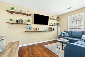 Un salon avec un canapé bleu, des étagères en bois ornées de plantes, un téléviseur à écran plat et un banc en bois.