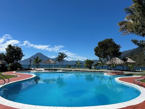 Una piscina con una terraza alrededor, palmeras y una montaña al fondo.