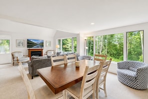 A modern living room with a wooden dining table, comfortable seating, a fireplace, and large windows offering a view of the forest.