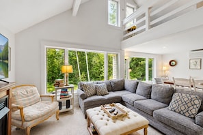 A living room with a sectional sofa, a chaise lounge, a coffee table, and a TV. There are books on a side table, and a window with a view of trees.