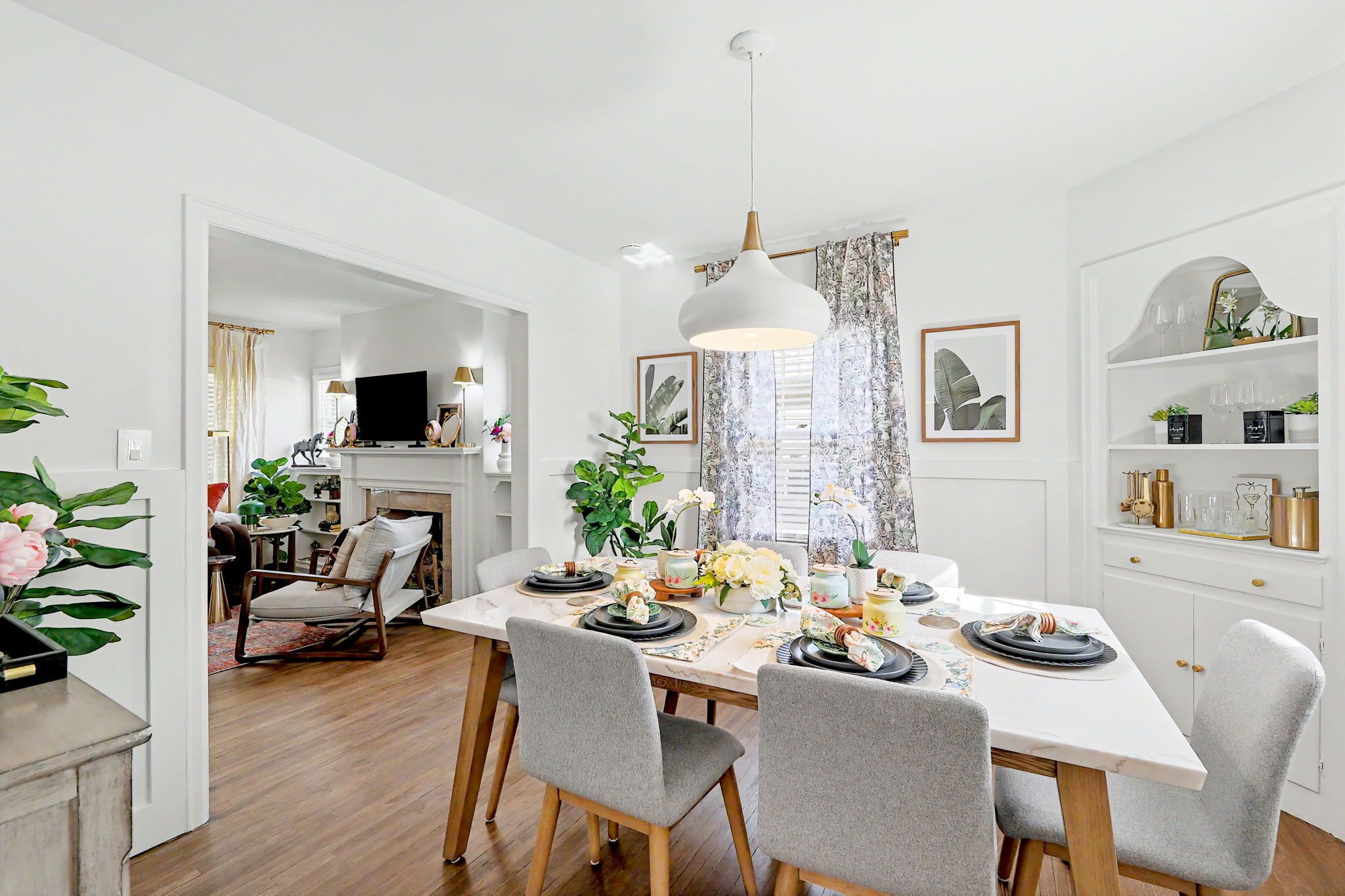 A dining area with a white table set for four, surrounded by grey chairs, a hanging light fixture, and a view of a living room with a television and plants.