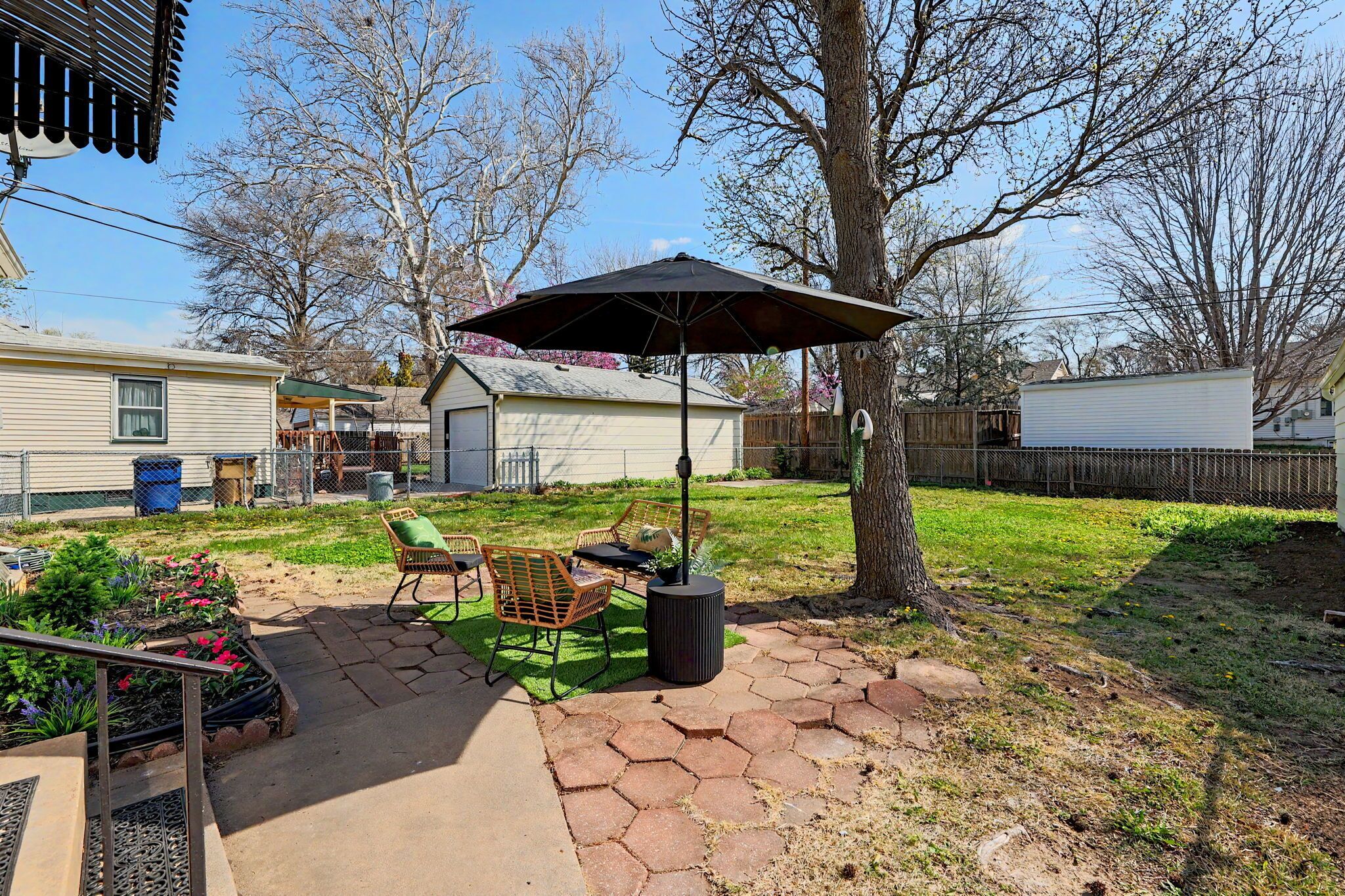 Un patio trasero con baldosas hexagonales, una mesa de metal, dos sillas, un cubo de basura y un árbol.