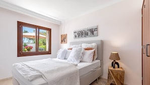 Un dormitorio con una cama, una ventana, una mesita de noche con una lámpara y una obra de arte colgada en la pared.