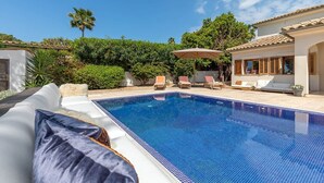 Una piscina con sillones de descanso y una sombrilla, rodeada de vegetación y un edificio con tejado de tejas.