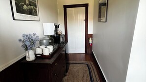 Een hal met een houten vloer, een donker vloerkleed, een koffiezetapparaat, een witte mok, een vaas met bloemen en een lijst met een foto aan de muur.