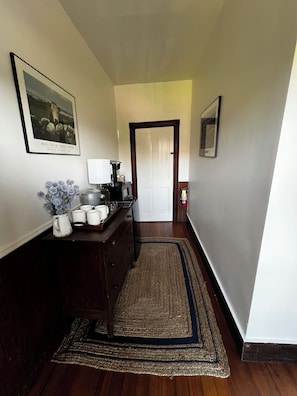 Un couloir avec un plancher en bois, un tapis sombre, une machine à café, une tasse blanche, un vase avec des fleurs et un tableau encadré accroché au mur.