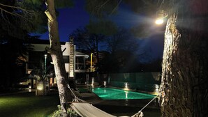 Una hamaca atada entre árboles, una piscina y un edificio moderno con grandes ventanales.