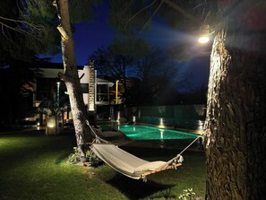 Eine Hängematte zwischen Bäumen, ein Schwimmbecken und ein modernes Gebäude mit großen Fenstern.
