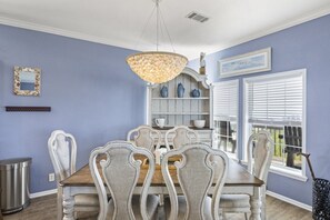 Uma sala de jantar com mesa e cadeiras de madeira, um lustre, um armário branco com vasos azuis e uma janela com persianas.