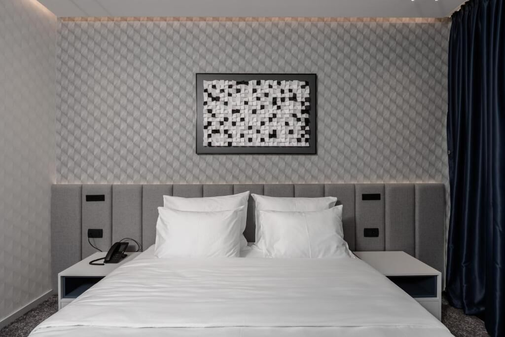 A modern hotel room with a neatly made bed, a headboard with storage compartments, a bedside table with a phone, and a patterned wall.