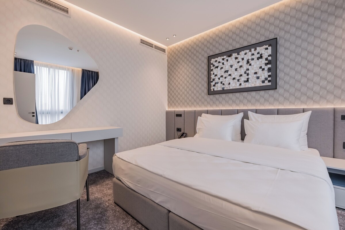 A modern hotel room with a large bed, a desk, a mirror, and a geometric wall art piece.