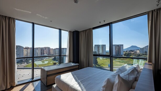 A modern hotel room with large windows, a bed, a bench, and a view of the cityscape. - Cube (Bar)