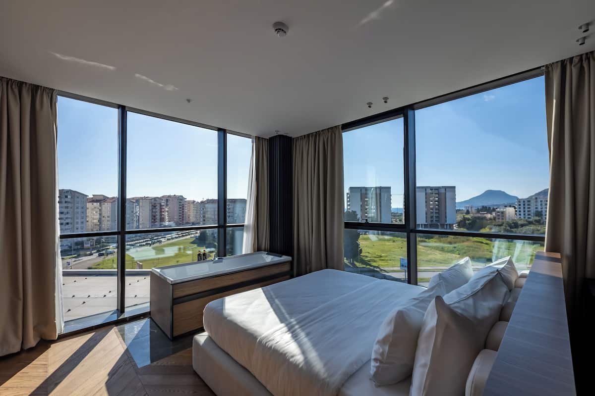 A modern hotel room with large windows, a bed, a bench, and a view of the cityscape.