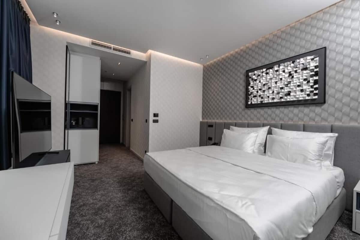 A modern hotel room with a large bed, a wardrobe, a desk, and a wall-mounted artwork.