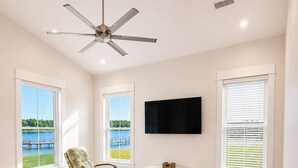 Una sala de estar con un sofá de cuero marrón, un sillón mecededor, un televisor de pantalla plana y ventanas amplias con vista a un cuerpo de agua.