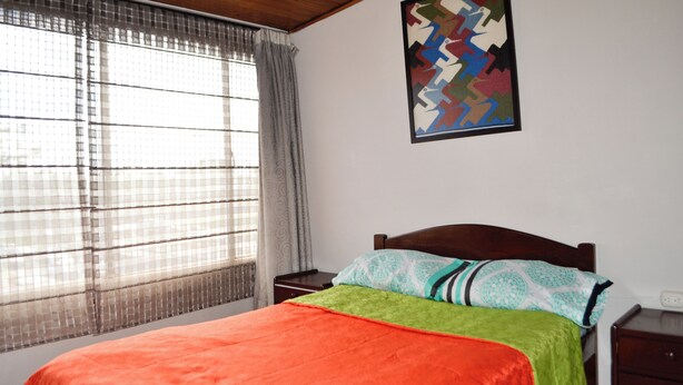 A bedroom with a bed, a window with curtains, a nightstand, and a wall-mounted artwork. - HOTEL DEL PUENTE BOGOTA (Bogotá)