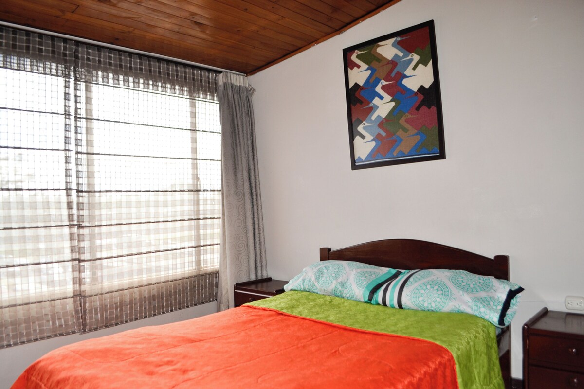 A bedroom with a bed, a window with curtains, a nightstand, and a wall-mounted artwork.