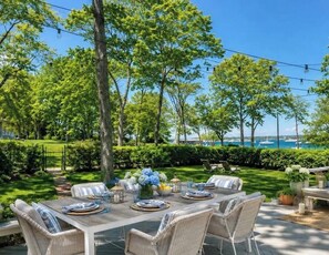 An outdoor dining area with a view of the sea, surrounded by trees and greenery. - Adorable Ram Island Water View Home & Guest Cottage with beach access (Shelter Island)