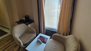 Ein Hotelzimmer mit Sitzgelegenheit, einem kleinen Tisch mit einer Zeitschrift, einer Kaffeemaschine und einem Fenster mit Vorhängen.