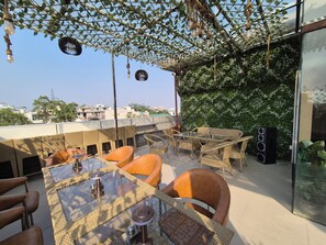 An outdoor seating area with wicker furniture and glass tables, surrounded by greenery and overlooking a cityscape. - OnlyStay Ginger Palace Sector 19 Noida (Noida)