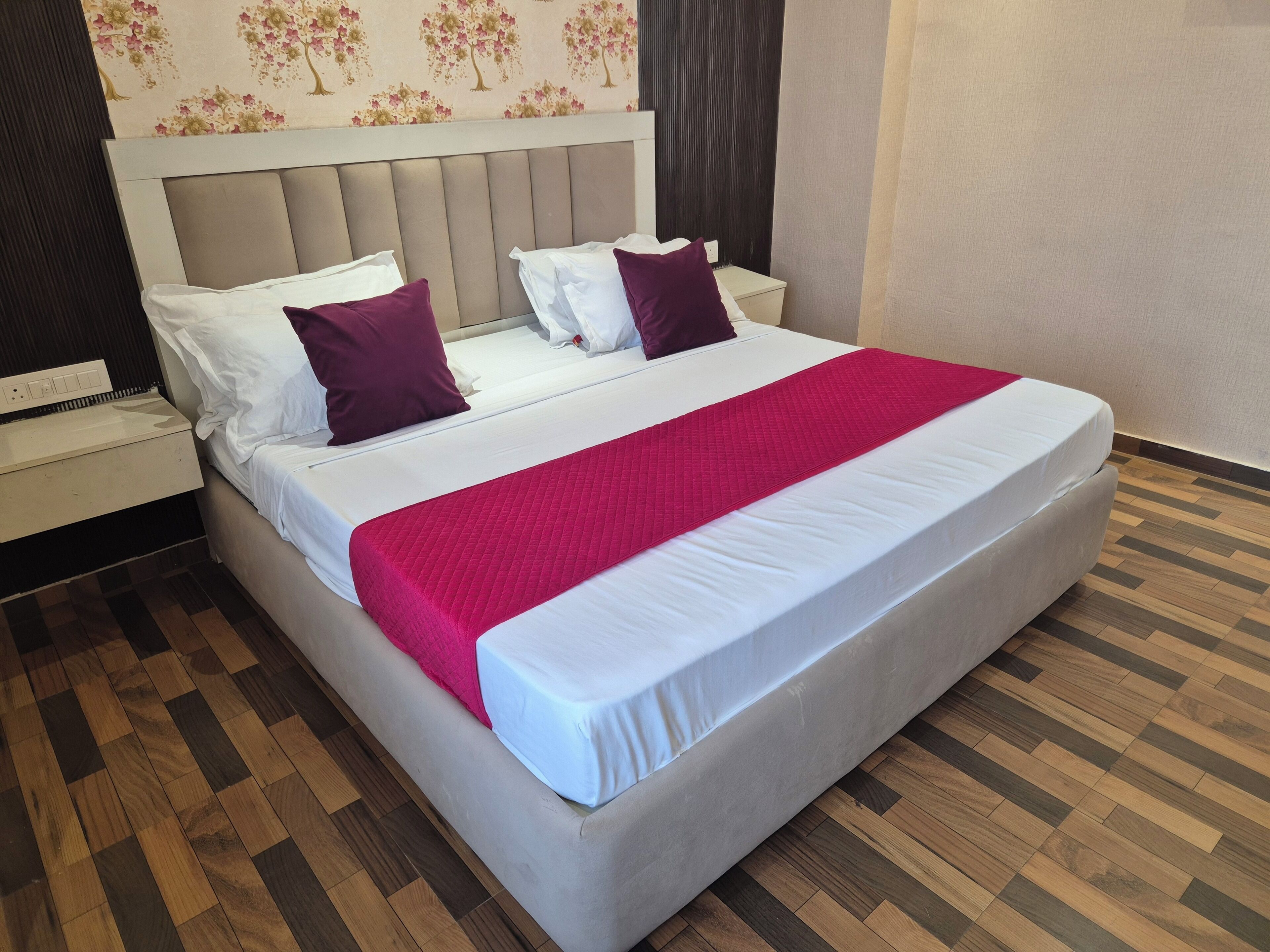 Una cama bien hecha con colchón blanco y manta rosa, cabecera con diseño de papel tapiz floral y suelo de parquet de madera.