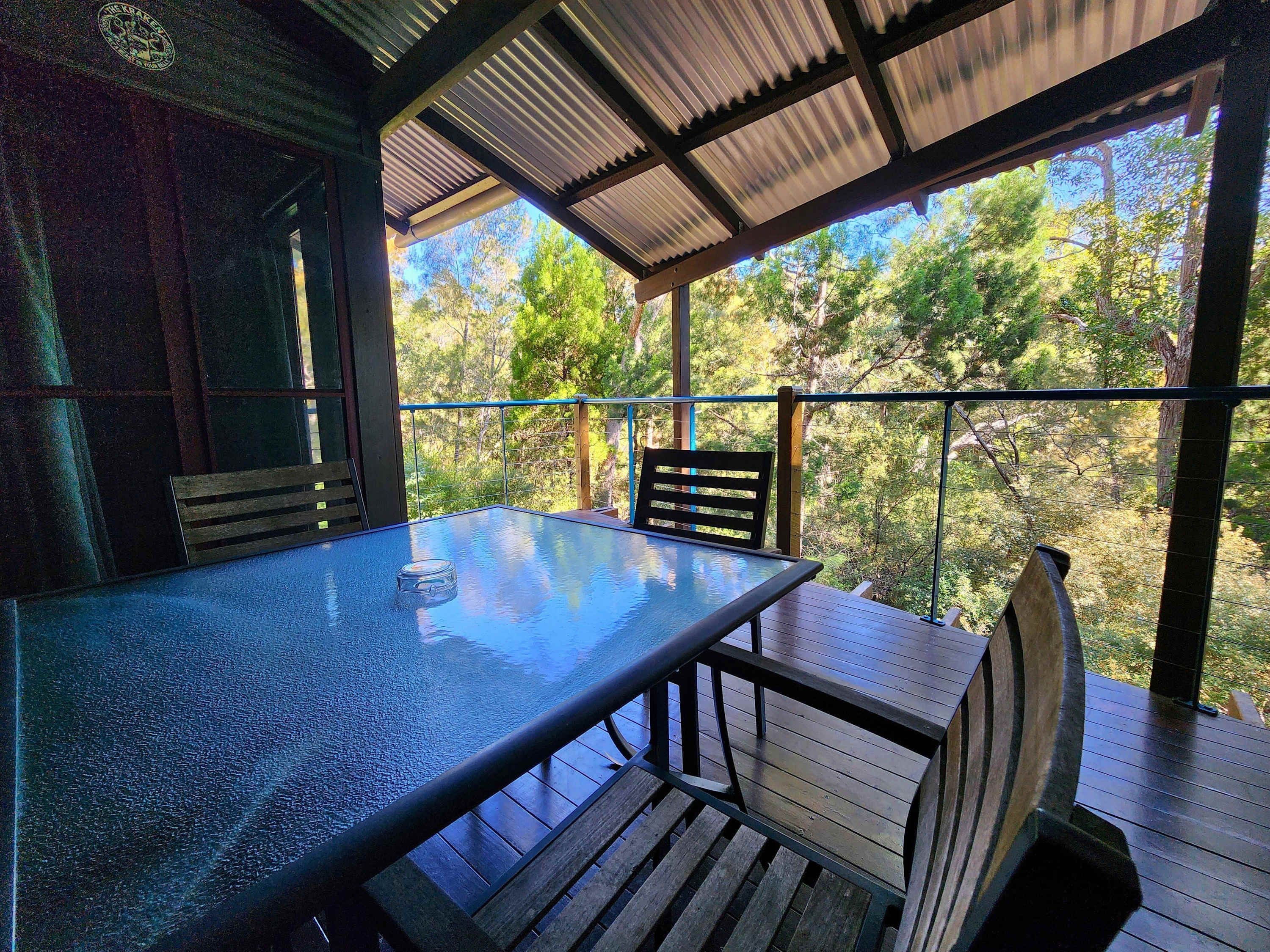 A covered outdoor seating area with a glass-top table and wooden chairs, surrounded by a metal railing.