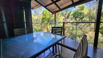 A covered outdoor seating area with a glass-top table and wooden chairs, surrounded by a metal railing.