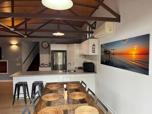 Een moderne keuken met een roestvrijstalen koelkast, een eethoek met een houten tafel en geweven placemats, en een schilderij van een zonsondergang aan de muur.