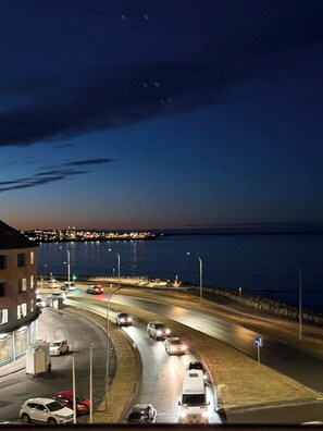 Vista nocturna d'una carretera costanera amb vehicles, un pont a la distància i un edifici a l'esquerra.