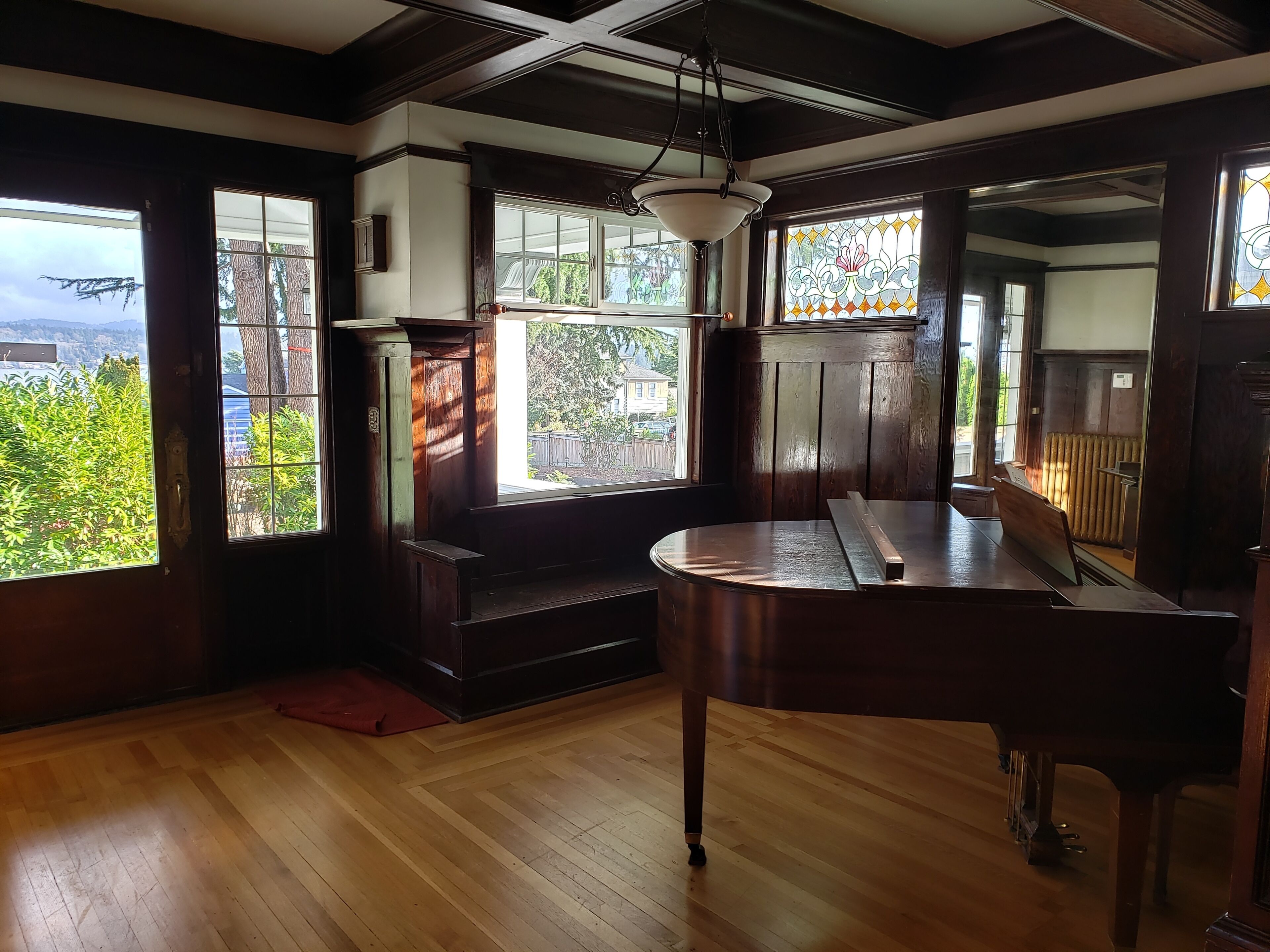 A room with a wooden floor, a piano, a bench, and large windows with a view of greenery.