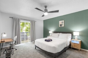 Habitación de hotel con cama, dos mesitas de noche con lámparas, un escritorio con silla, ventilador de techo y vistas a través de una ventana con cortinas.