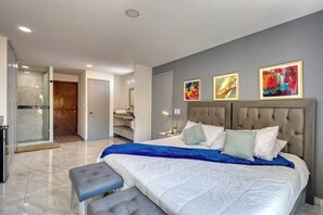 A bedroom with a large bed, a bench, a shower area, and three framed artworks on the wall. - Hotel Suite With Ocean-view Rooftop Infinity Pool, Balconies & AC (Cabo San Lucas)